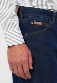 Jeans de mezclilla azul oscuro con un parche de cuero, remaches de cobre y costuras en contraste, combinados con un puño de camisa blanca a rayas.