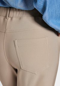 Beige stretch stof broek met een achterzak, met een gladde textuur en een verborgen tailleband, gedeeltelijk bedekt door een blauwe blouse.