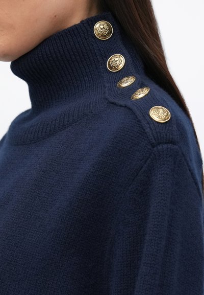 Marineblauwe trui met hoge kraag, gemaakt van textuur breiwerk, met vijf gouden decoratieve knopen langs de schouder voor extra detail.