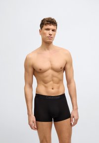 Ευδιάθετος άντρας χωρίς πουκάμισο, φορώντας μαύρα εσώρουχα Calvin Klein, στέκεται μπροστά σε ένα απλό ανοιχτό γκρι φόντο.