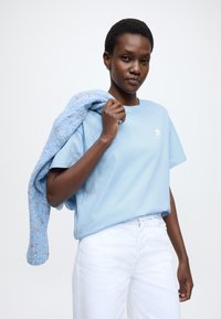 Ανοιχτό μπλε βαμβακερό t-shirt με άνετη εφαρμογή, που φέρει ένα μικρό λευκό λογότυπο. Μια υφασμάτινη μπλε μπλούζα με πολύχρωμες πιτσιλιές κρεμασμένη πάνω στον ώμο.