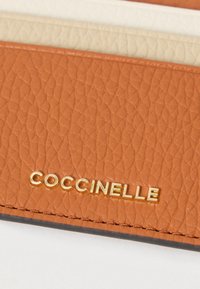 Καφέ πορτοφόλι από δερμάτινη υφή με το ανάγλυφο όνομα της μάρκας «Coccinelle» σε χρυσά γράμματα. Λείος εσωτερικός χώρος με ανοιχτόχρωμες αποχρώσεις.