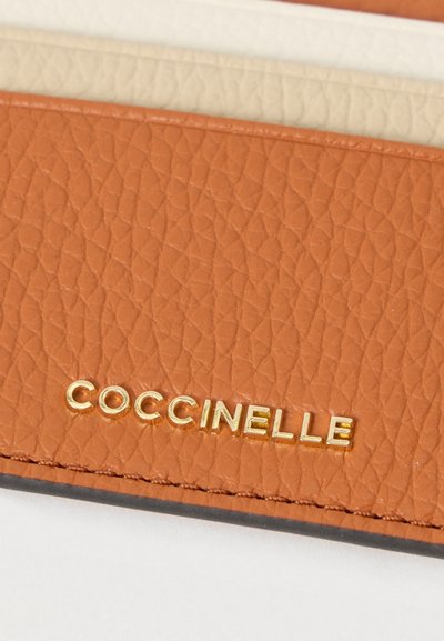 Portefeuille en cuir marron texturé avec le nom de la marque "Coccinelle" embossé en lettres dorées. Intérieur lisse avec des teintes plus claires.