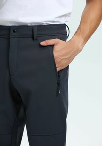 Hand delvis inne i den dragkedjade fickan på mörkblå byxor som bärs med en vit skjorta.