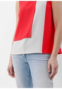 Rood en wit mouwloos top met gelaagd ontwerp, gemaakt van gladde stof, gecombineerd met lichtblauwe jeans. Close-up van handen op de taille.