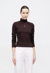 Bordowy prążkowany sweter z golfem i długimi rękawami, z małą naszywką z logo na piersi, uzupełniony jasnoróżowymi spodniami.