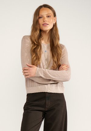 Donna con lunghi capelli mossi in piedi con le braccia incrociate, indossa un cardigan beige lavorato a maglia con bottoni multicolore e pantaloni neri su sfondo semplice.