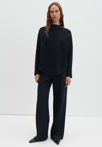 Maglione a coste blu navy con collo alto, abbinato a pantaloni a gamba larga e décolleté con punta a punta, su uno sfondo chiaro e semplice.