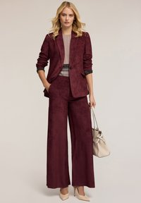Traje de terciopelo burdeos con chaqueta a medida y pantalones de pierna ancha. Combina con una blusa clara y un bolso crema. Presenta una textura suave.