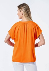 Orangenes Kurzarm-Bluse mit entspannter Passform, Faltendetail am Ausschnitt und glatter Textur. Getragen mit weißen Hosen.