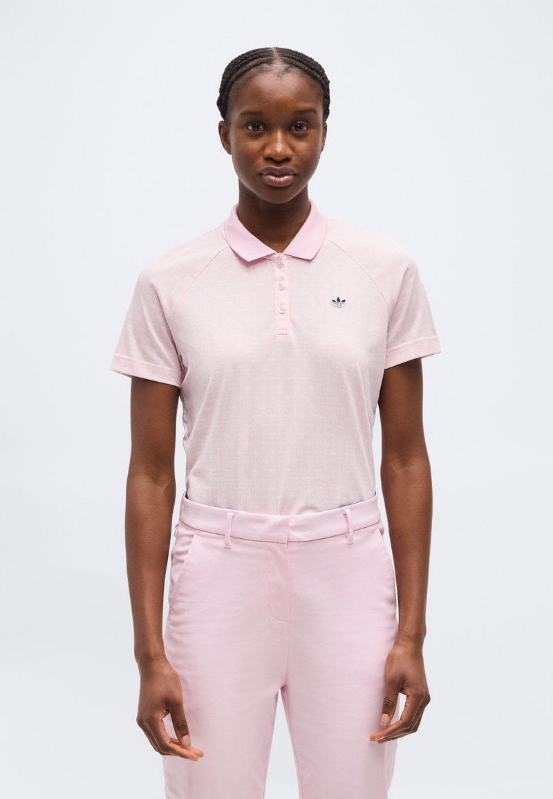 Νεαρή γυναίκα με πλεξούδες, ντυμένη με ροζ κοντομάνικο πόλο της Adidas και παντελόνι που ταιριάζει, στέκεται μπροστά σε ένα απλό λευκό φόντο.