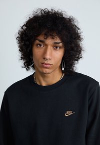 Čierna mikina Nike s malým oranžovým logom na hrudi, okrúhlym výstrihom a štruktúrovanou textíliou. Viditeľné kučeravé vlasy a piercings.