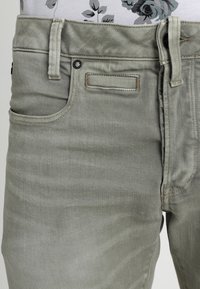 Jean en denim vert clair avec une texture lisse, dotée de poches avant, de passants de ceinture et d'accents en laiton. Taille plate avec des détails de couture minimaux.