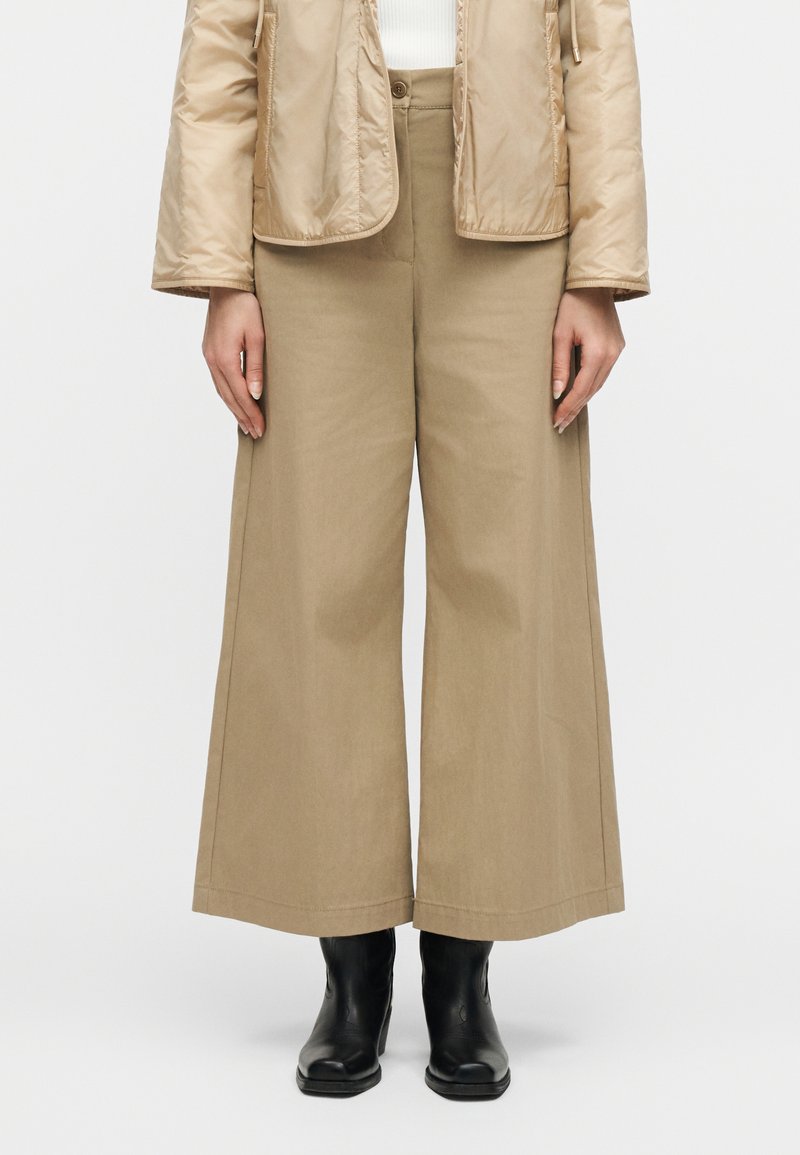 Μπεζ παντελόνι με φαρδιά γραμμή συ νδυασμένο με μαύρες μποτάκια έως τον αστράγαλο και ανοιχτό μπεζ καπιτονέ μπουφάν, απεικονιζόμενο από μπροστά από τη μέση μέχρι τα πόδια.