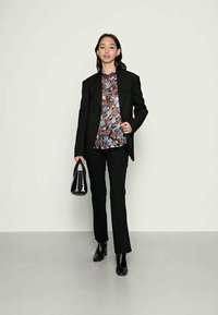 Blazer preto ajustado sobre uma blusa floral multicolorida, combinada com calças pretas e botas ankle, segurando uma pequena mala preta.