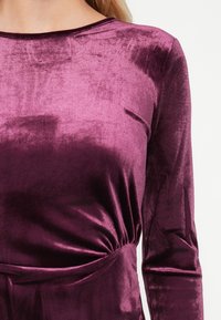 Gros plan d'une personne portant un haut en velours violet profond à manches longues avec un détail froncé à la taille sur un fond clair.
