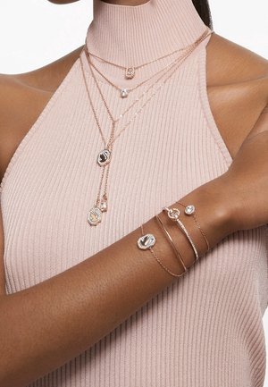 Femme portant des colliers superposés en or avec de petits pendentifs et des bracelets assortis en or, vêtue d'un débardeur côtelé rose pâle.