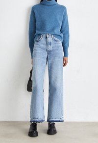 Pull col roulé bleu clair, coupe ample, associé à un jean en denim clair taille haute, jambes larges avec ourlet effiloché, et bottes noires épaisses.