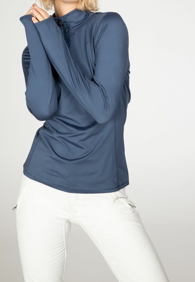 Haut de sport à manches longues bleu marine en tissu lisse et extensible ; dispose d'un col haut et de trous pour les pouces. Associé à un pantalon slim blanc.