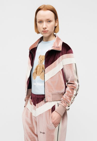 Βελούδινο φόρμας σε ροζ και μπορντό με κρεμ ρίγες. Διαθέτει ζακέτα με φερμουάρ και ελαστική μέση. Κάτω από αυτή, ένα ανοιχτό μπλε μπλουζάκι με στάμπα αρκουδάκι.