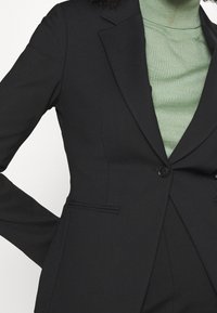 Persona che indossa un blazer nero attillato su un maglione a collo alto a coste di un verde chiaro, mentre posa con una mano sulla vita.