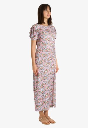 Donna in piedi che indossa un abito maxi rosa a fiori con maniche a sbuffo e sandali beige, leggermente rivolta verso destra su uno sfondo bianco semplice.