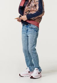 Bambino in jeans azzurri chiari, sneakers bianche e rosa della Nike e una giacca a motivi con dettagli blu navy e rosa, in piedi su uno sfondo bianco uniforme.