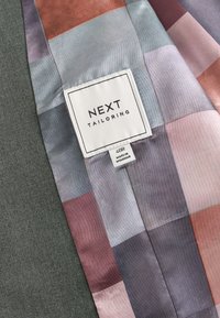 Beklædningsstof med et flerfarvet geometrisk mønster og et påsyet mærke med teksten "NEXT TAILORING", størrelse 40R, fremstillet i Myanmar.