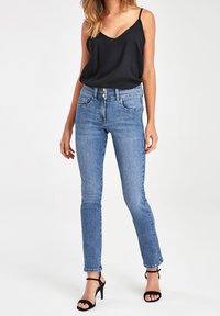 Top negro sin mangas, jeans ajustados azules con cierre de botón y cinco bolsillos, finalizados con sandalias de tacón negro. Tejido suave y diseño clásico.