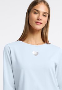 Lichtblauwe shirt met lange mouwen, gemaakt van zachte stof, met een zilver metallic hartontwerp op de borst. Eenvoudige ronde halslijn.