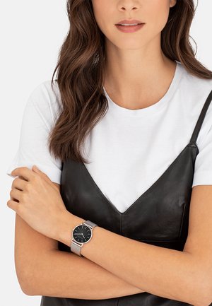 Robe sans manches en cuir noir sur un t-shirt blanc, arborant une montre avec un bracelet en maille argentée, un cadran foncé et des marqueurs d'heure minimalistes.