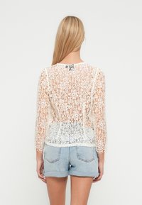 Κορυφή από δαντέλα με μακριά μανίκια, κρεμ χρώμα, με λουλουδάτα μοτίβα και ημιδιαφανή σχεδίαση, φορεμένη πάνω από ανοιχτόχρωμα τζιν σορτς.