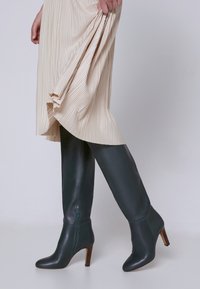 Femme levant une jupe plissée beige pour montrer des bottes en cuir noir jusqu'aux genoux avec des talons en bois et une fermeture éclair sur le côté, sur fond uni.