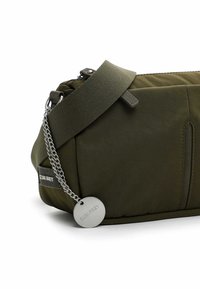 Borsa di tessuto verde oliva con texture imbottita, tracolla regolabile, accento in catena metallica e un'etichetta circolare argentata. Dotata di chiusura con zip.