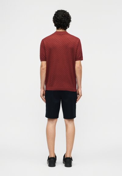 Άνδρας, με την πλάτη, φοράει υφασμάτινη κοντομάνικη πουκάμισο πόλο μπορντό χρώματος, μαύρη βερμούδα και μαύρα παπούτσια σε απλό λευκό φόντο.