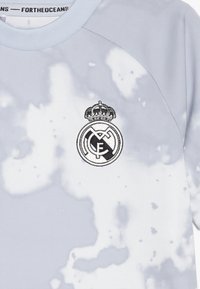 Svetle šedý sportovní svetr s texturovaným, abstraktním vzorem; na hrudi má černě vyšitý znak Real Madrid.