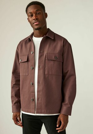 Bordeauxkleurig overhemd met knoopjes, voorzien van twee voorzakken, een textuurstof en een puntkraag, gedragen over een wit T-shirt, gecombineerd met zwarte broek.