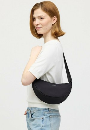 Jonge vrouw met schouderlang rood haar, draagt een wit topje met korte mouwen en lichtblauwe spijkerbroek, en draagt een zwarte halvemaanvormige schoudertas.