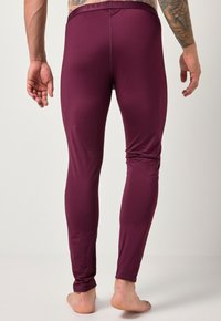 Uomo con un braccio tatuato che indossa leggings aderenti color bordeaux, in piedi a piedi nudi contro uno sfondo chiaro e neutro, vista di spalle.