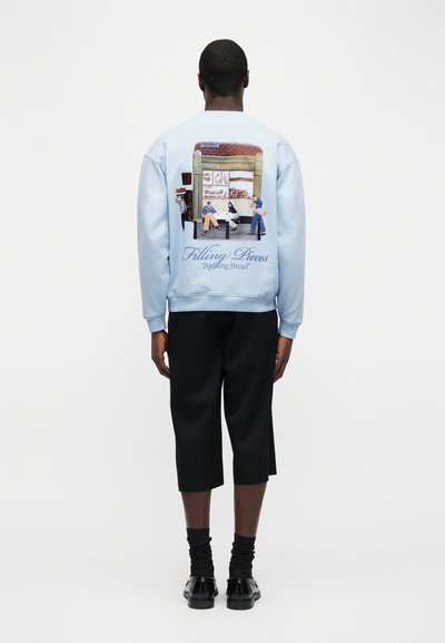 Άνδρας που στέκεται με την πλάτη, φοράει ανοιχτό μπλε φούτερ με γραφικό σχέδιο ενός φούρνου και ανθρώπων που κάθονται και περπατούν στην πλάτη.