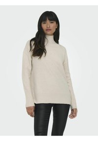 Pull à col montant beige avec des poignets et un ourlet côtelés, fabriqué en un tissu tricoté doux, porté avec un pantalon en cuir noir.