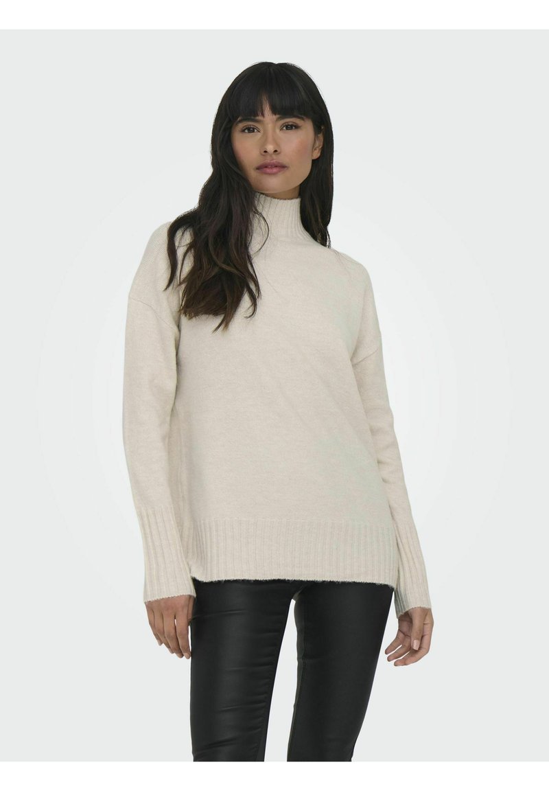 Pull à col montant beige avec des poignets et un ourlet côtelés, fabriqué en un tissu tricoté doux, porté avec un pantalon en cuir noir.