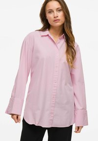 Šviesiai rožinė medvilninė marškinėlių palaidinė su apykakle, ilgomis rankovėmis ir plačiomis rankogaliais, turinti laisvą siluetą.