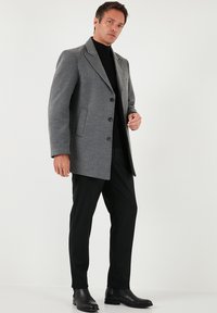 Sivi volneni plašč z zarezanimi ovratniki in štirimi črnimi gumbi, kombiniran s črnimi hlačami in usnjenimi gležnarji. Gladek tekstura, krojen po meri.