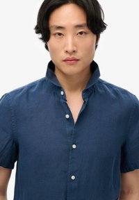 Joven con cabello negro que lleva una camisa de manga corta azul oscuro con botones y cuello abierto, sobre un fondo blanco liso.