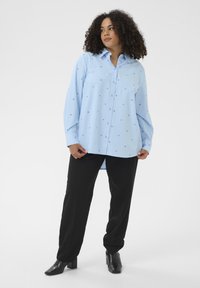 Chemise bleu clair à boutons avec petits motifs de cœurs bleus. Dotée d'un col et de manches longues, assortie à un pantalon noir et des bottines à talons.