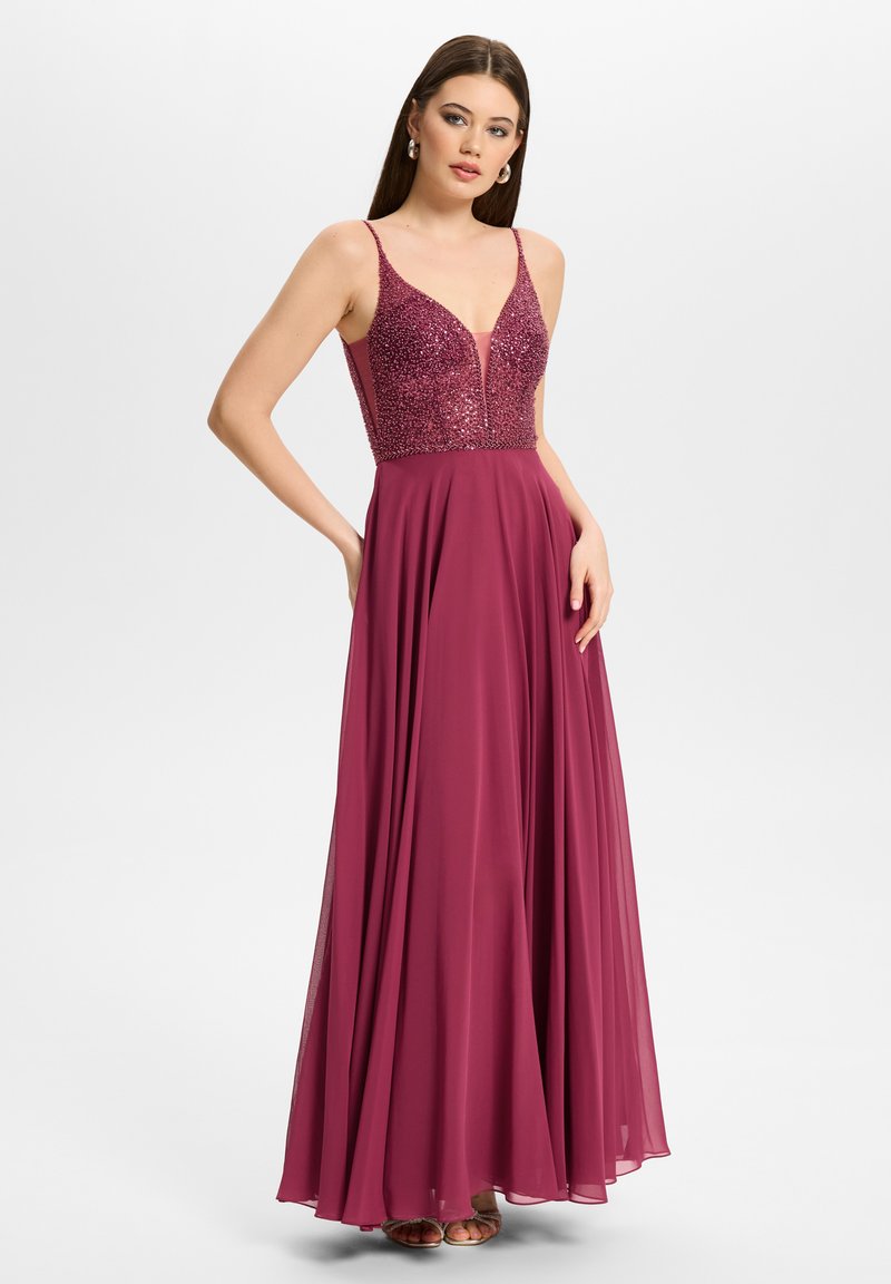 Joven mujer con un vestido largo, sin mangas, de color rosa oscuro, con corsé bordado con cuentas y falda fluida, posando sobre un fondo liso.