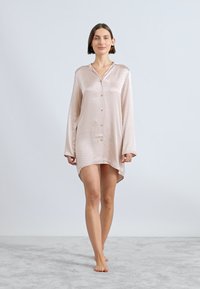 Rochie cămașă din satin roz pal, cu un model texturat, cu deschidere pe butoane, mâneci lungi, care se termină la mijlocul coapsei și are un tiv curbat.