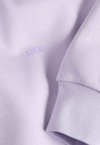 Light lavender sweatshirt fabric with a smooth texture features a subtle "JXXX" print in the same color. The sleeve cuff is tightly woven.