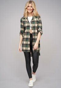 Camicia a maniche lunghe a plaid verde e beige, abbottonata, indossata sopra un top corto bianco, abbinata a leggings neri e sneakers bianche.